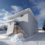 This is a colour photograph of a two-storey white residential building (numbered 44) taken on a sunny winter day. The building has white horizontal siding and several windows. A wooden exterior staircase with railings leads up to the main entrance, which includes a small covered porch. The roof and surrounding ground are heavily covered in deep snow. The sky is bright blue with scattered white clouds.