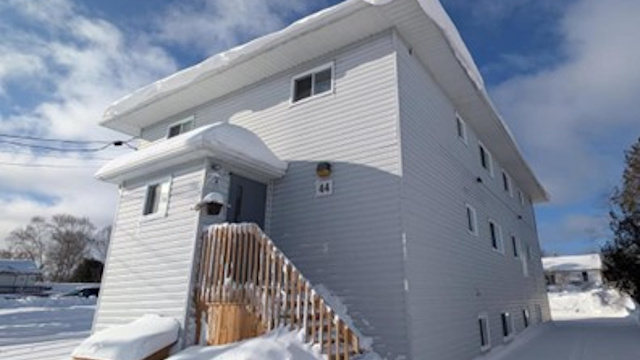 Vue extérieure d’un immeuble résidentiel de deux étages en hiver. Le bâtiment est recouvert de parement clair et possède plusieurs fenêtres réparties sur les façades visibles. Un petit escalier en bois mène à une entrée située sur le côté gauche, avec une rampe et de la neige accumulée autour des marches. De la neige recouvre également le sol et les abords du bâtiment. Le ciel est partiellement nuageux, laissant entrer la lumière naturelle. D’autres bâtiments sont visibles à l’arrière‑plan, indiquant un secteur résidentiel.