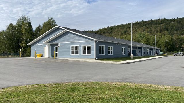 Vue extérieure d’un bâtiment de plain‑pied de forme allongée, situé dans un secteur dégagé. Le bâtiment est recouvert d’un revêtement bleu pâle avec des fenêtres blanches réparties le long des façades. Une entrée principale avec un auvent est visible à l’extrémité gauche. Un vaste stationnement asphalté entoure le bâtiment, avec des lampadaires et quelques zones gazonnées au premier plan. Des arbres et une colline boisée apparaissent en arrière‑plan, sous un ciel partiellement nuageux.